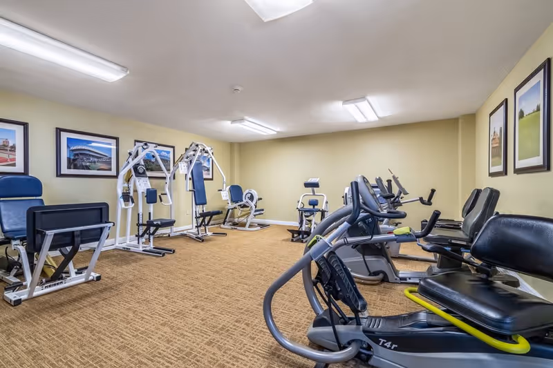 A fitness room with various exercise equipment including stationary bikes, weight machines, and rowing machines. The room has beige walls, carpeted flooring, and framed pictures hanging on the walls.