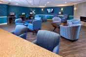 Spacious senior living lounge with multiple upholstered armchairs and side tables, computer stations along the back wall, and a TV on a teal-accented wall.