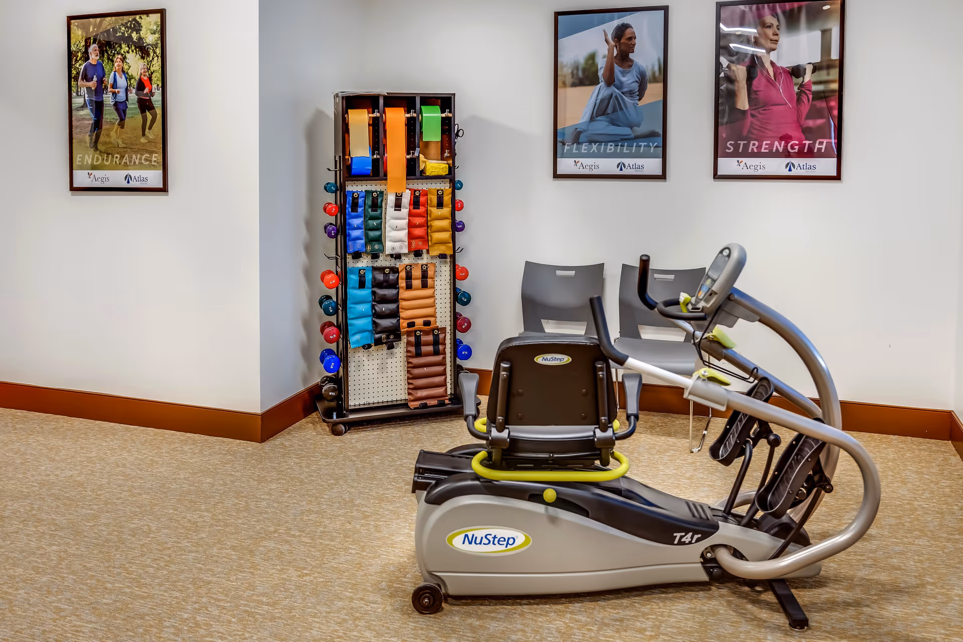 A small exercise room with a NuStep T4r recumbent cross trainer, two gray chairs, and a rack holding colorful resistance bands and small dumbbells. Three framed posters on the wall depict people exercising with the words Endurance, Flexibility, and Strength.