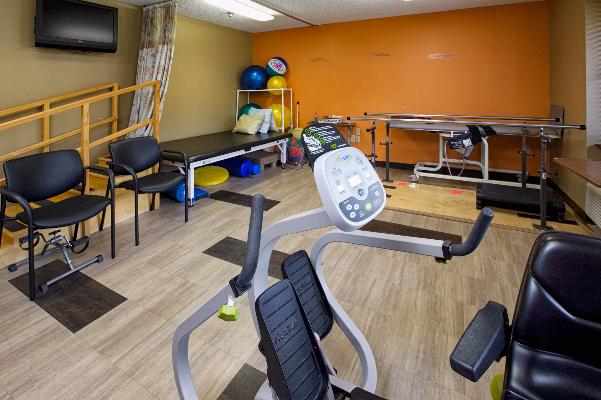Interior therapy/rehab room with exercise equipment, parallel bars, a treatment table, exercise balls and chairs.
