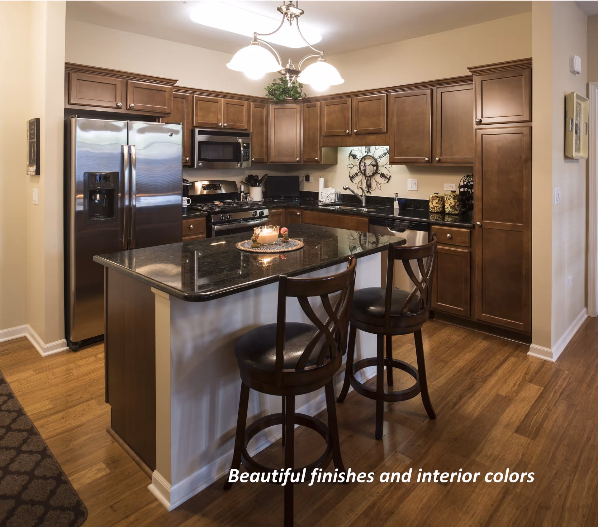 A modern kitchen with wooden cabinets, stainless steel refrigerator, microwave, and stove. There is a black granite countertop island with two wooden bar stools. A decorative clock is mounted on the wall above the sink. The floor is wooden, and a light fixture with three lamps hangs from the ceiling.