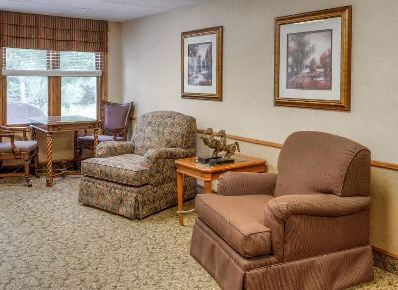 A cozy sitting area with two armchairs, one upholstered in a floral pattern and the other in a solid brown fabric, separated by a wooden side table with a decorative horse sculpture. Behind the chairs are two framed landscape paintings on a beige wall. To the left, there is a window with blinds and a valance, and a small wooden table with two chairs.