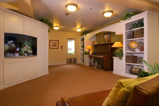 A cozy senior living common room with a piano, built-in bookshelves, an aquarium, lamps, and seating.