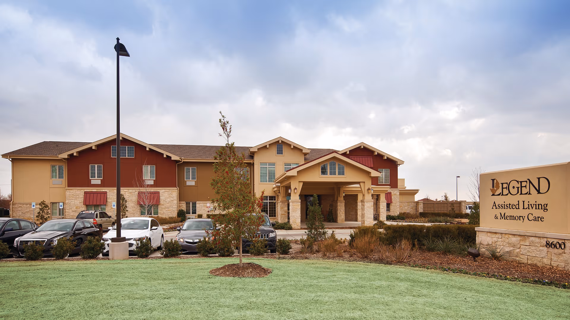 Exterior view of Legend Assisted Living & Memory Care facility in Fort Worth, showing a two-story building with a covered entrance, several parked cars, a green lawn, and a sign with the facility's name.