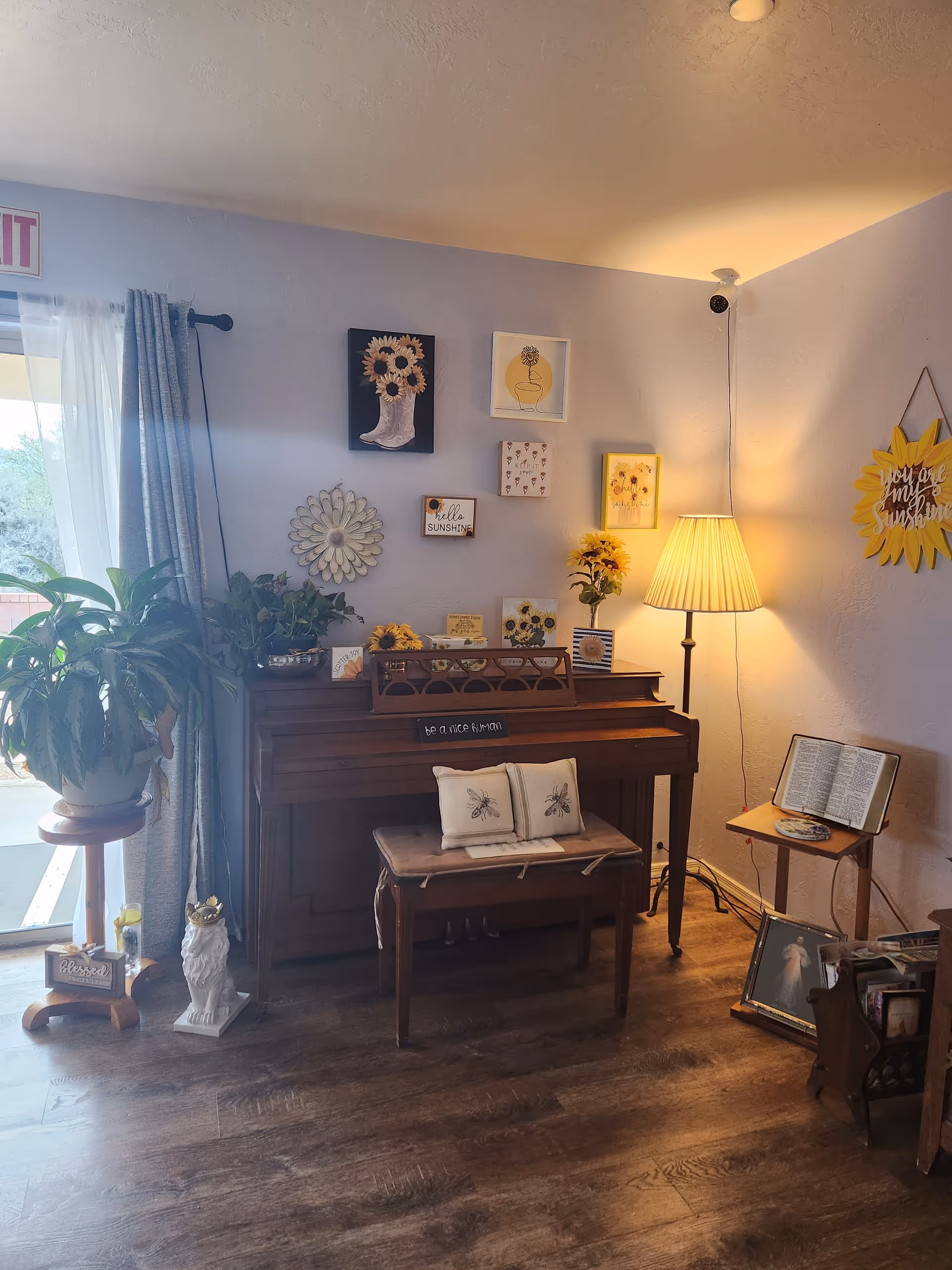 Cozy corner of a living room with a wooden piano adorned with sunflower-themed decorations and a small bench with two bee-embroidered pillows. A floor lamp with a pleated shade illuminates the area. Nearby, a small table holds an open book, and a plant stands on a wooden stool next to a window with sheer curtains. Various sunflower and floral wall art decorate the light purple walls.