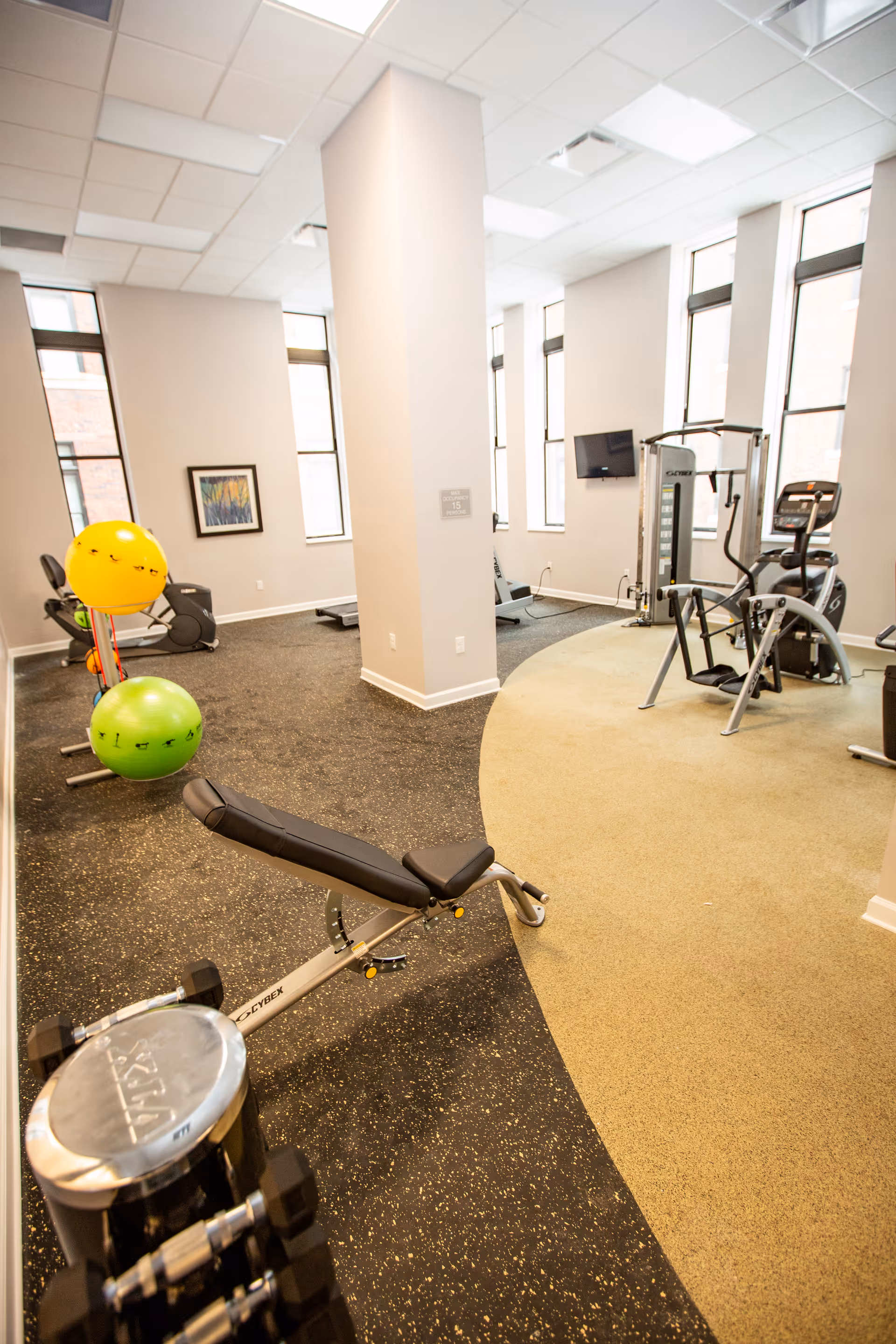 A bright fitness room with exercise equipment including a weight bench, dumbbells, exercise balls, and cardio machines. The room has large windows letting in natural light, light-colored walls, and a mix of carpet and rubber flooring.