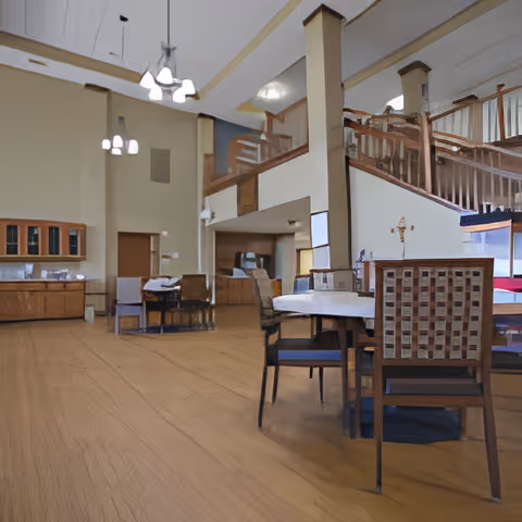 Spacious interior room with wooden flooring and furniture, including a round table with chairs in the foreground and a staircase with wooden railing leading to an upper level. The room has high ceilings with hanging light fixtures and a cabinet along one wall.
