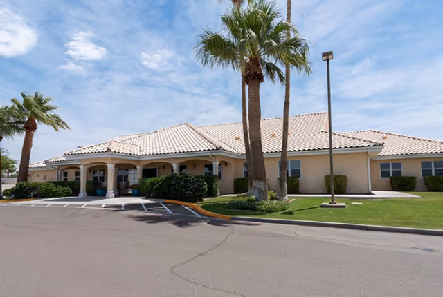 Single-story senior living building front with a tiled roof, covered entrance, palm trees, and a driveway.