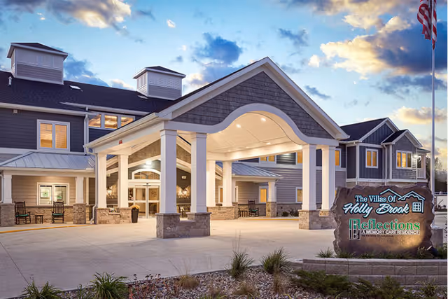 Exterior view of Villas of Holly Brook Assisted Living & Memory Care building at dusk, featuring a large covered entrance with white pillars, stone accents, and multiple windows. The sky is partly cloudy with a warm glow from the setting sun. A stone sign with the facility's name and logo is visible in the foreground near landscaped plants.