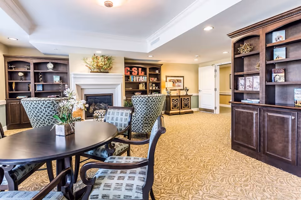 A cozy senior living common area featuring a dark oval table with four patterned chairs, two armchairs with a geometric design, built-in dark wood bookshelves filled with decorative items and books, a fireplace with a floral arrangement on the mantel, and a beige patterned carpet. The room is softly lit with recessed lighting and a ceiling light fixture.