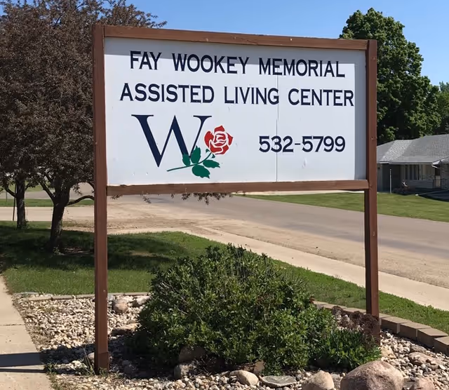 A roadside sign for Fay Wookey Memorial Assisted Living Center with a 'W' and rose logo and a phone number on a grassy lawn.
