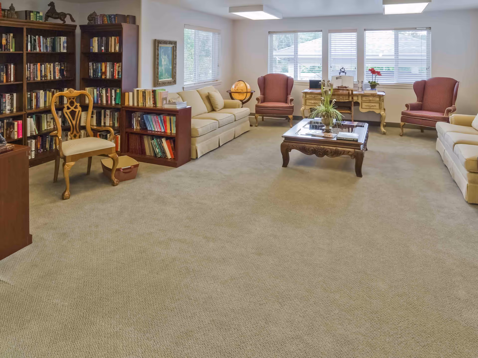 A bright communal living room with sofas, armchairs, bookshelves, a coffee table and a desk beneath windows.