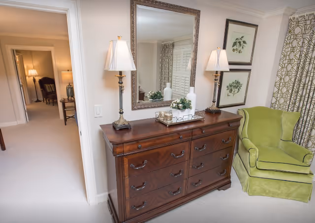 A cozy interior corner of a retirement community room featuring a wooden dresser with multiple drawers, two decorative table lamps with white shades, a large framed mirror above the dresser, and a green upholstered armchair next to patterned curtains. The room has light-colored walls and carpet, with a hallway visible in the background.