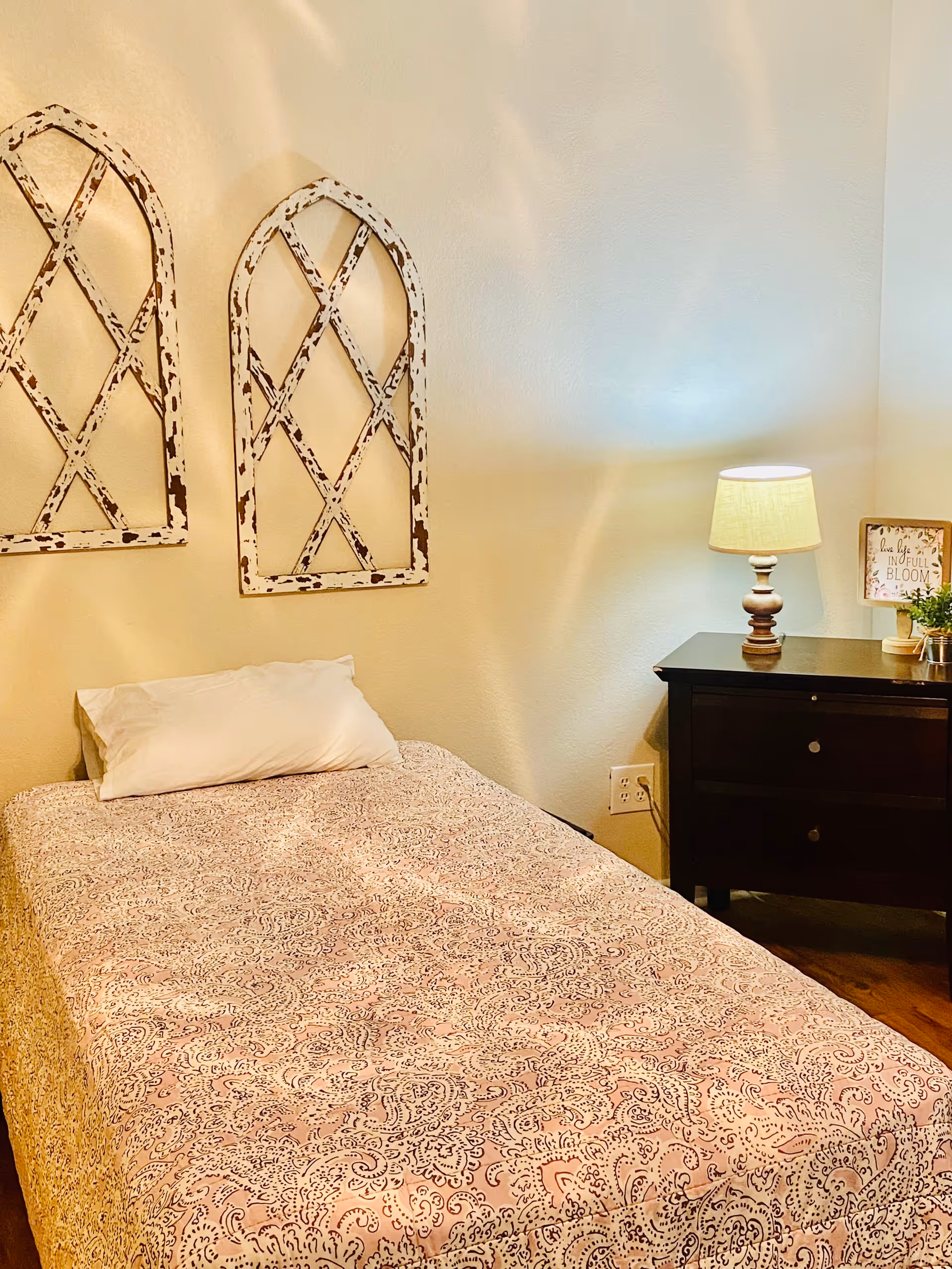 A cozy bedroom with a single bed covered in a patterned bedspread and a white pillow. Above the bed are two decorative rustic wooden wall hangings with a distressed white finish. Next to the bed is a dark wooden nightstand with two drawers, a lit table lamp, a small potted plant, and a framed sign that reads 'Live life in full bloom'. The walls are painted a light beige color.