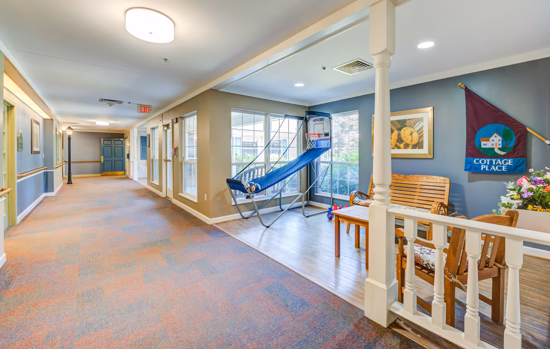 A bright hallway in a senior living facility with carpeted floors and beige walls. On the right side, there is a small seating area with wooden chairs, a bench, a table with flowers, and a basketball arcade game. Large windows let in natural light. A flag on the wall reads 'Cottage Place'.