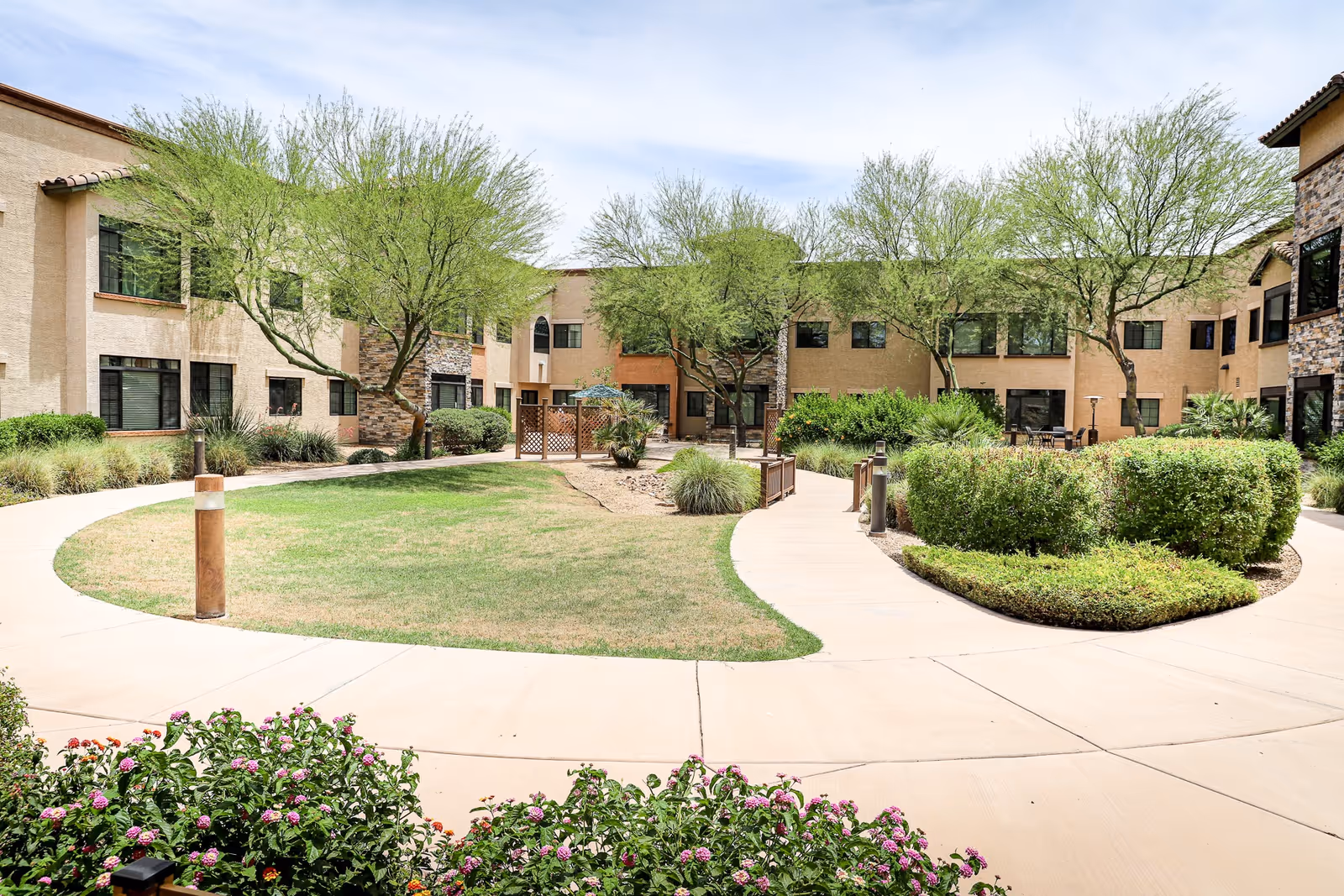 Outdoor courtyard area at Orchard Pointe at Surprise featuring a circular walkway surrounding a grassy lawn with trees and shrubs, bordered by a two-story building with multiple windows.