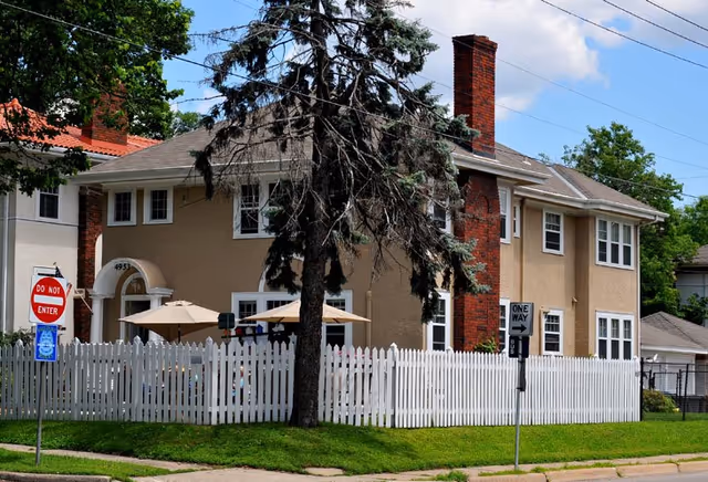 Exterior view of a two-story beige building with white trim, a red brick chimney, and a white picket fence surrounding a small yard with patio umbrellas. There are street signs including a 'Do Not Enter' and a 'One Way' sign near the sidewalk.