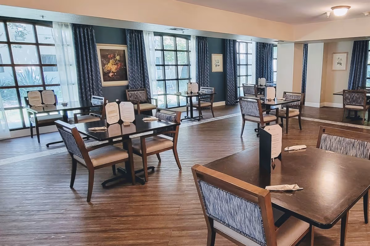 Spacious dining room with multiple square tables and chairs arranged near large windows and curtains.