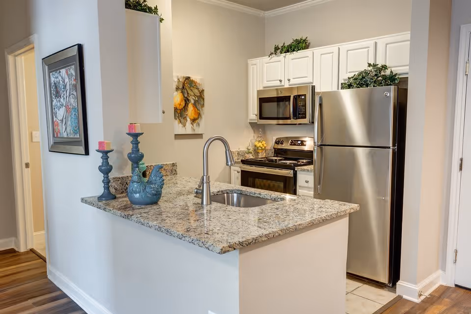 Modern kitchen with granite countertops, stainless steel refrigerator, stove, and microwave. The kitchen island features a sink and decorative items including blue candlesticks and a ceramic rooster. White cabinets and light-colored walls complete the space.