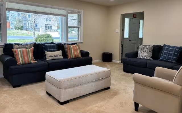 A cozy living room with two dark blue sofas adorned with colorful and patterned throw pillows, a beige armchair, and a large beige ottoman in the center. There is a large window behind one sofa letting in natural light and showing a view of a residential street outside. The walls are painted light beige and the carpet is a matching beige color.