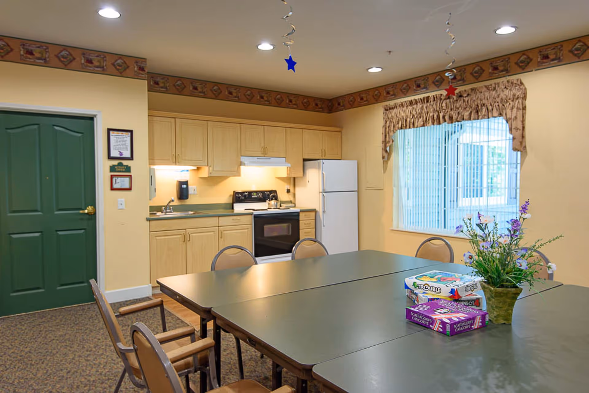 A well-lit room with a kitchen area featuring light wood cabinets, a white refrigerator, a stove, and a sink. In front of the kitchen is a large table with several chairs around it. On the table are board games and a vase with flowers. The room has a window with vertical blinds and a decorative valance, and the walls are painted a soft yellow with a patterned border near the ceiling.