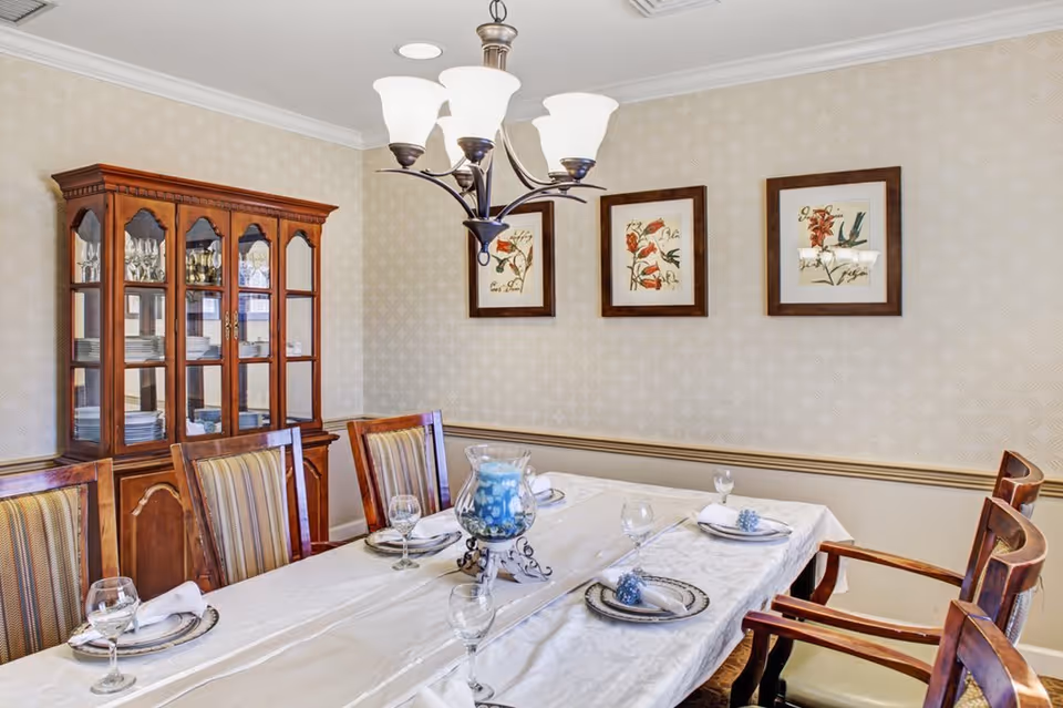 A formal dining room with a long table covered with a white tablecloth, set with plates, napkins, and wine glasses. There are six wooden chairs with striped upholstery around the table. A wooden china cabinet filled with glassware and plates is against the wall. Three framed floral artworks hang on the patterned wallpaper. A chandelier with five lights hangs above the table.