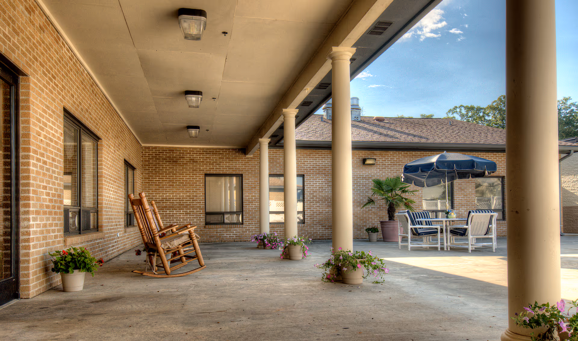 Covered outdoor patio area with brick walls, several columns, two wooden rocking chairs, potted plants, and a table with chairs and a blue umbrella in the background.