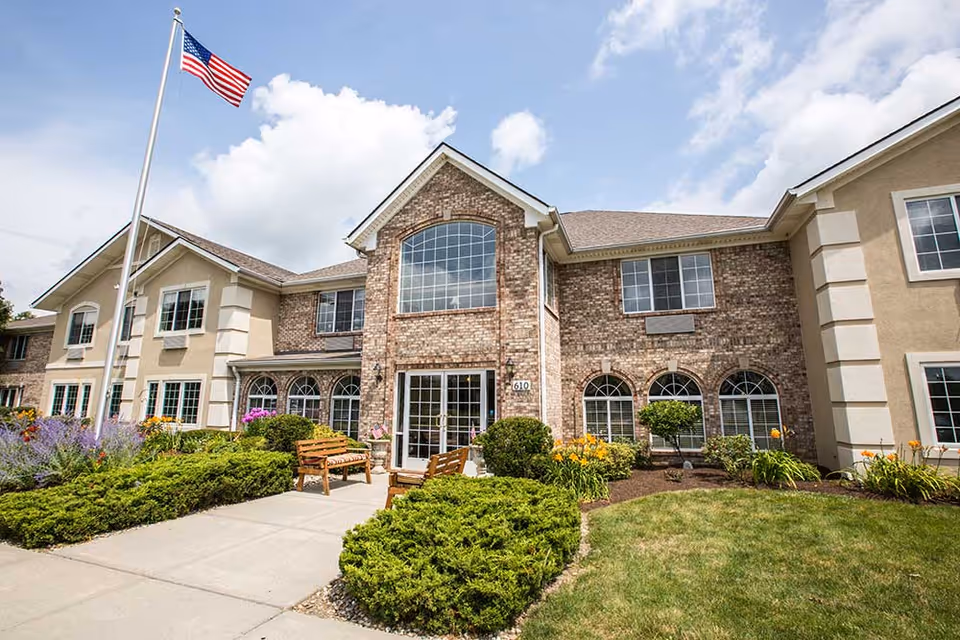 Exterior view of Crown Pointe senior living facility showing a two-story building with brick and beige siding, large windows, a flagpole with an American flag, landscaped bushes, flowers, and benches along a concrete walkway under a partly cloudy sky.