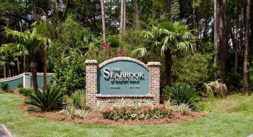 A landscaped entrance sign for The Seabrook of Hilton Head, surrounded by green grass, palm trees, and other plants with a wooded area in the background.