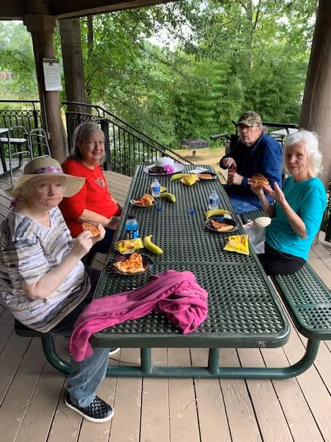 Four elderly people sitting around a green outdoor picnic table on a covered porch, eating pizza and bananas. The table has water bottles, napkins, and plates with food. Trees and greenery are visible in the background.