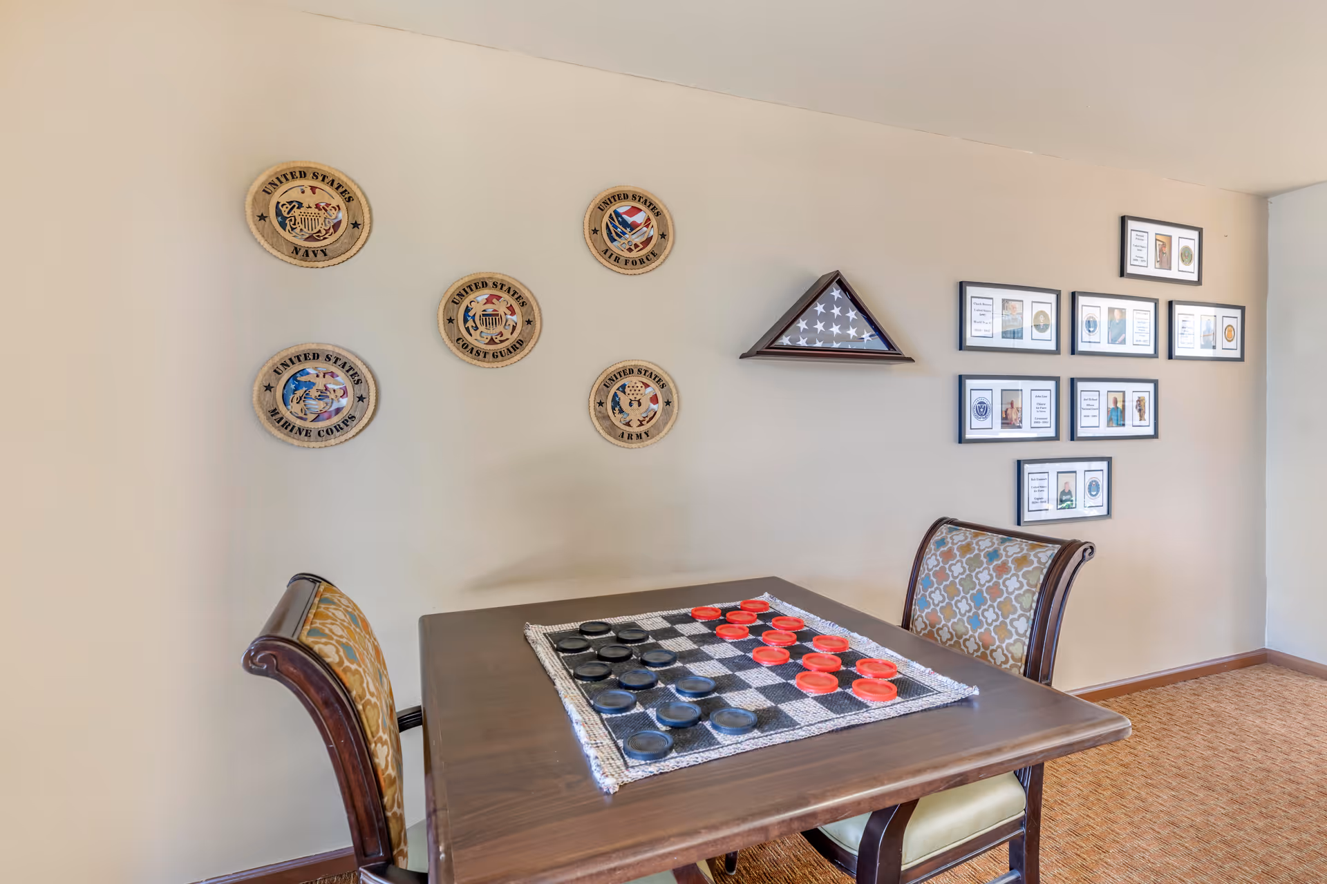 A small wooden table with a checkers game set up, surrounded by two upholstered chairs with patterned fabric. The wall behind the table displays plaques representing different branches of the United States military and a folded American flag in a triangular display case, along with framed pictures and documents.