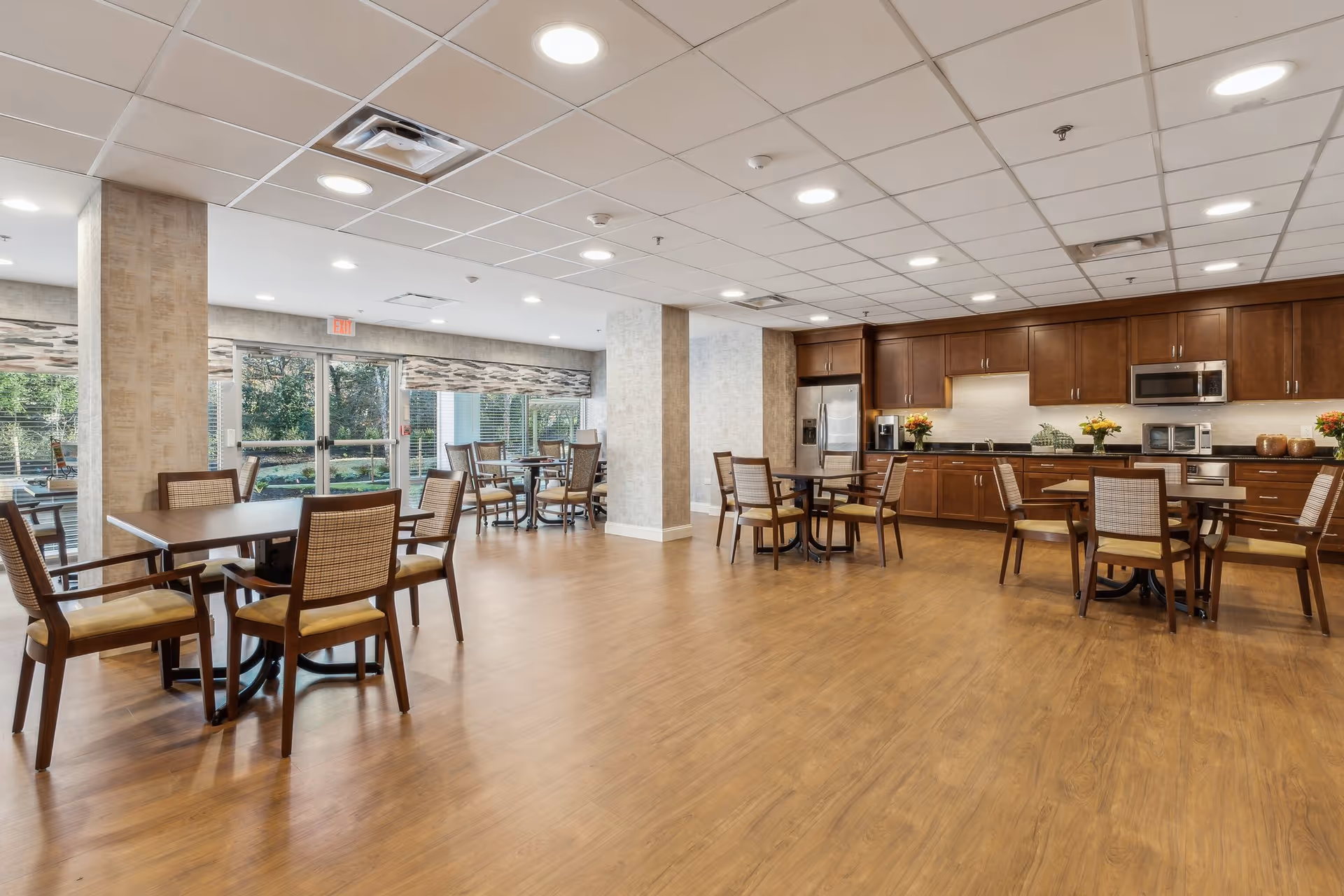 Communal dining room with multiple tables and chairs and a kitchen area with wooden cabinets and stainless appliances.