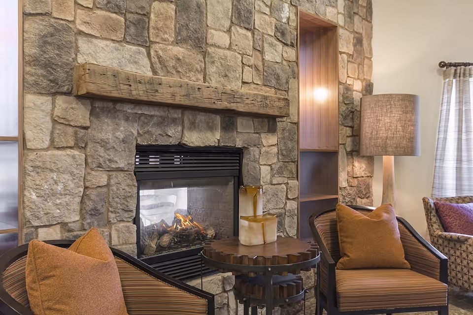 Cozy living room area with a stone fireplace featuring a wooden mantel. Two cushioned armchairs with orange pillows are positioned around a small round wooden table with decorative items. A floor lamp with a beige shade and a window with curtains are visible in the background.