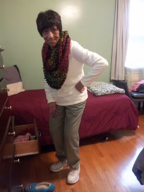 An older woman wearing a colorful scarf stands in a small bedroom next to a bed with a red bedspread.