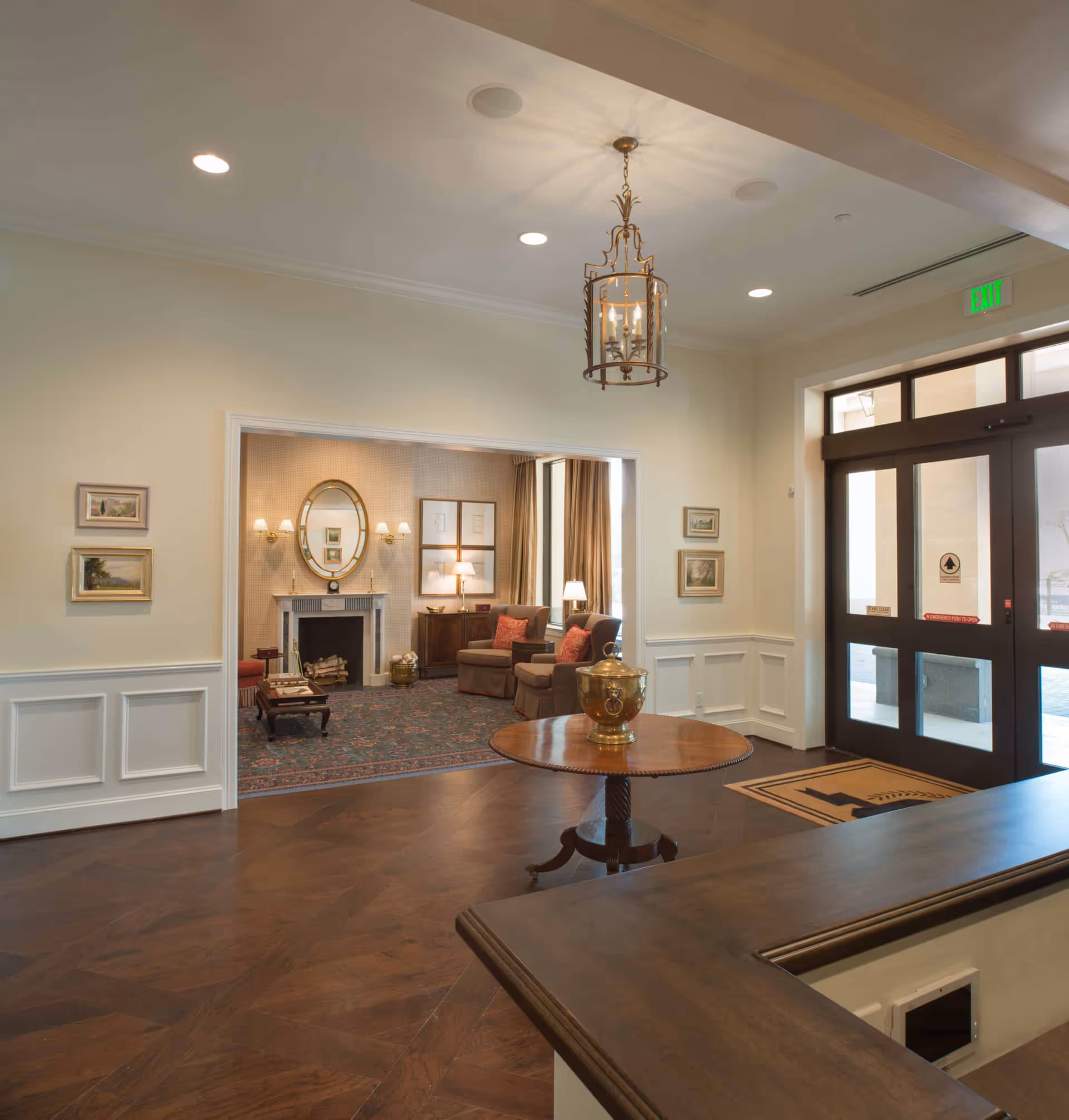 Bright entry lobby with wood floors, a round center table and chandelier leading to a cozy seating area with a fireplace and front glass doors.