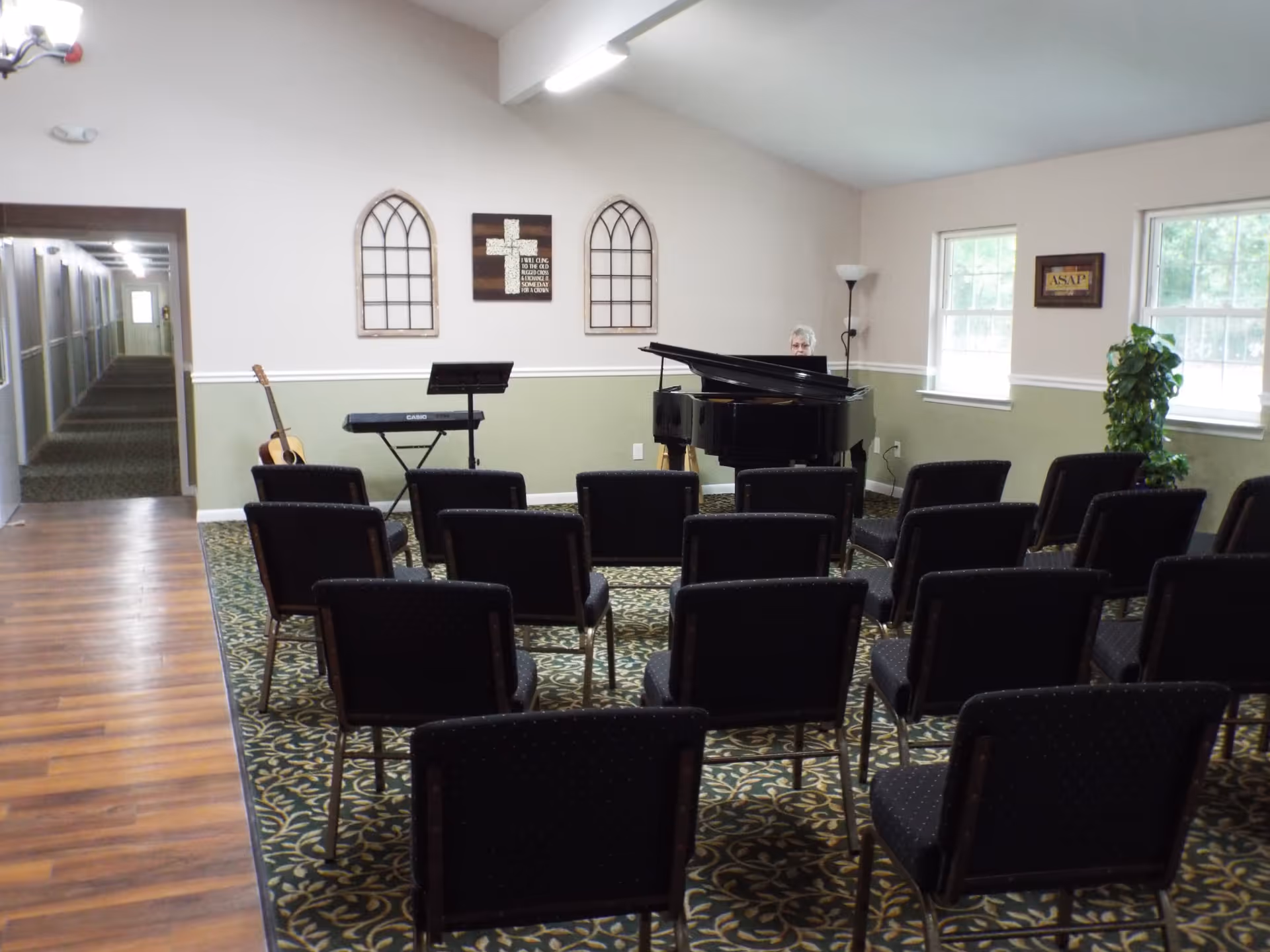 A small music room with rows of black chairs facing a grand piano where an elderly person is seated. There is also a keyboard on a stand and an acoustic guitar resting against the wall. The walls are decorated with two window-shaped wall hangings and a cross with a religious quote. Two windows let in natural light, and a green plant is placed near one window.
