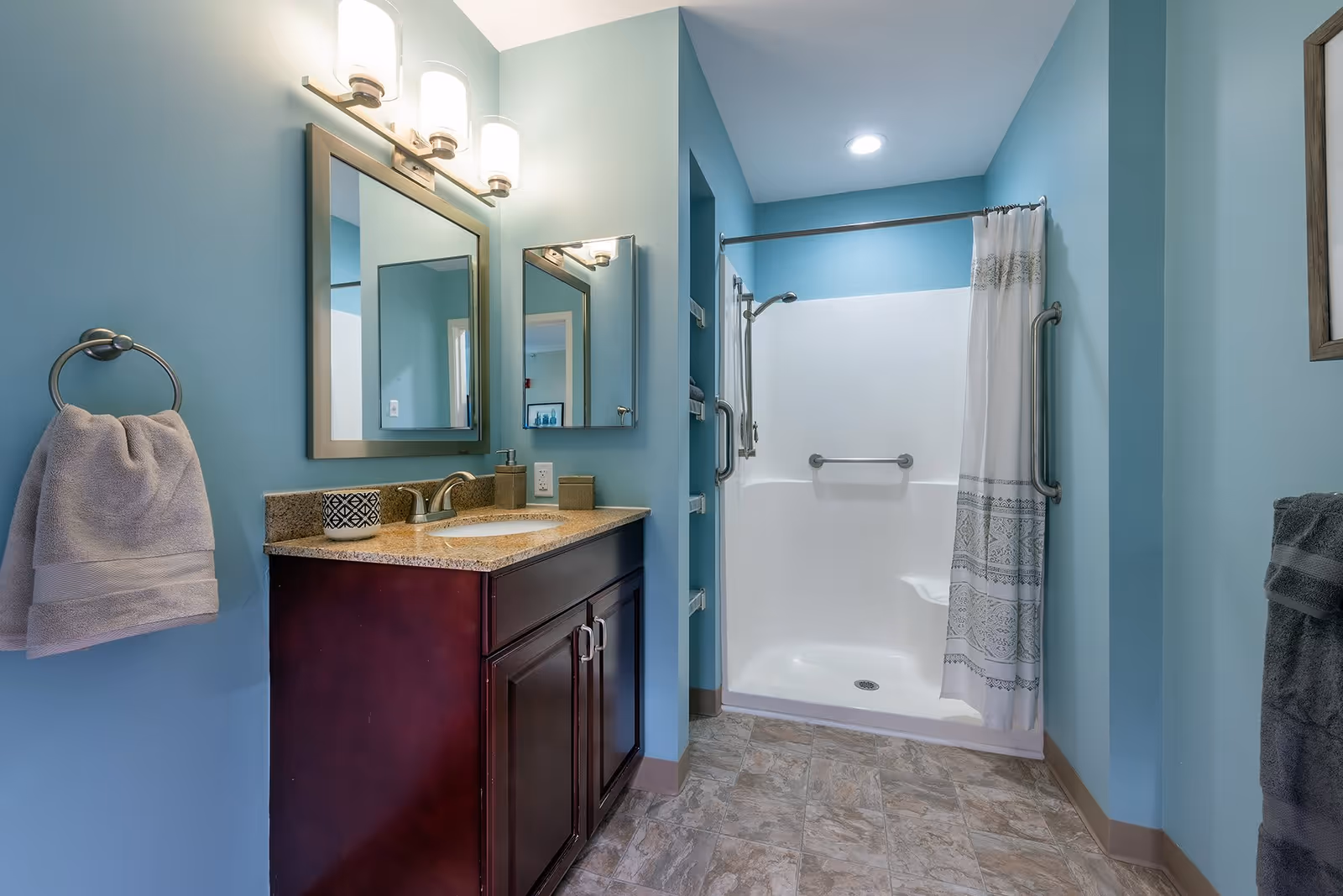 A clean bathroom with light blue walls featuring a dark wood vanity with a granite countertop and an undermount sink. Above the sink is a large mirror with three light fixtures. To the right is a walk-in shower with grab bars and a white shower curtain with decorative patterns. The floor has a tile pattern in neutral tones.