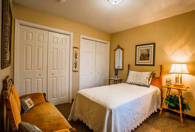 Cozy bedroom with a single bed covered in a white bedspread, bedside table and lamp, closet doors and a small sofa.