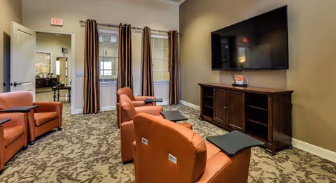 A cozy living room area with four orange armchairs arranged facing a large flat-screen TV mounted on a beige wall. Below the TV is a dark wooden media console. The room has patterned carpet flooring, beige walls, and three windows with brown and beige striped curtains. An open door leads to another room with additional furniture visible.