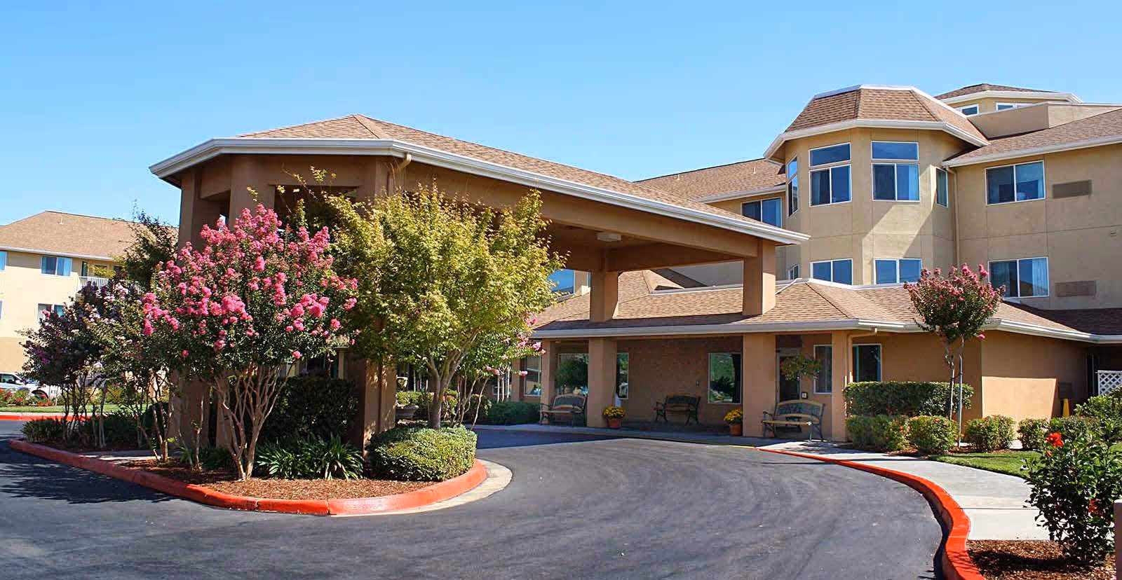 Exterior view of Solstice Senior Living at Lodi showing the entrance driveway with a covered drop-off area, surrounded by landscaped bushes and flowering trees under a clear blue sky.