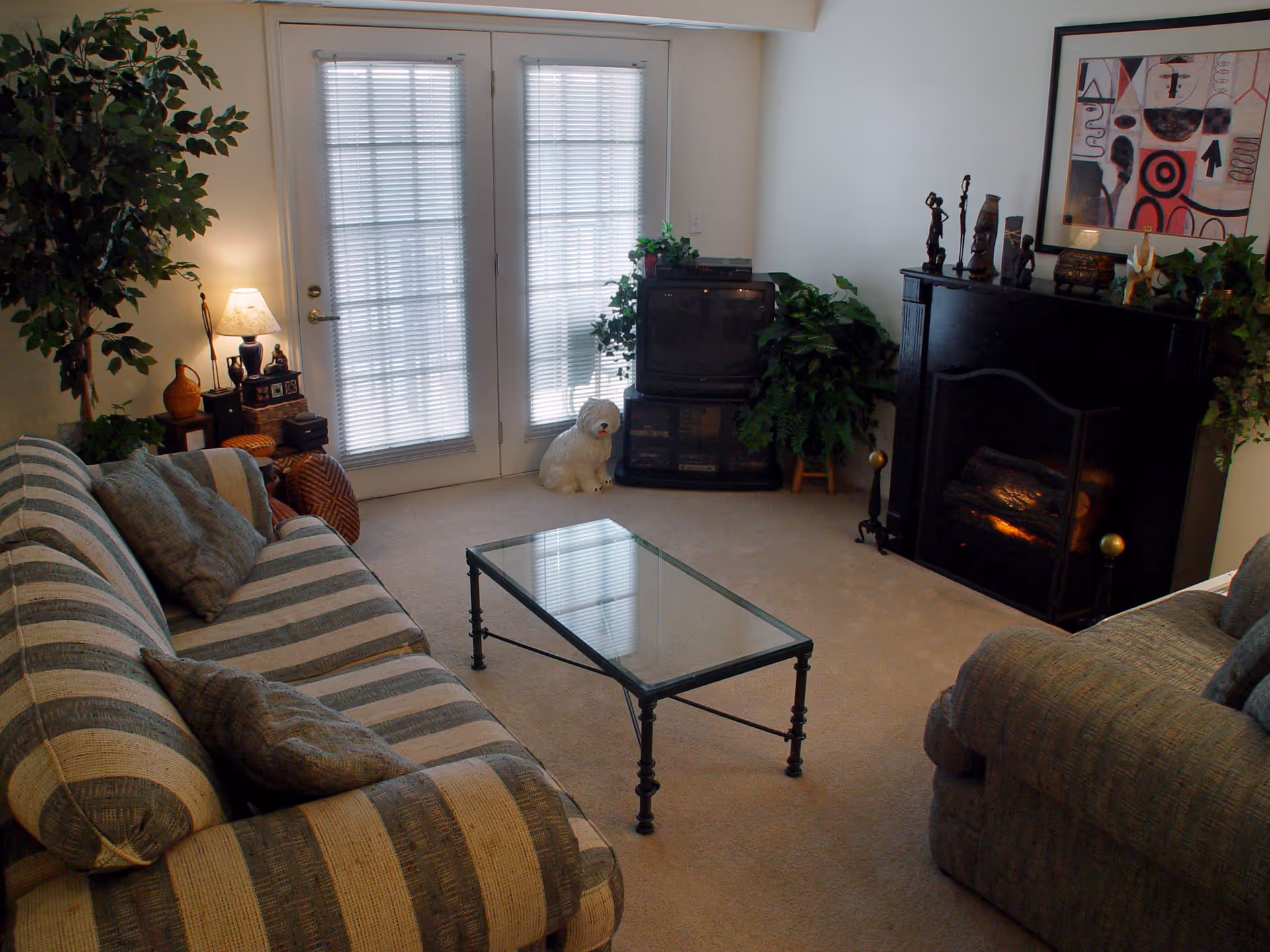 A cozy living room with a striped sofa and armchair, a glass coffee table, a black fireplace with decorative items on the mantel, a TV on a stand, potted plants, a lamp on a side table, and French doors with blinds letting in natural light.