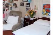 A cozy nursing home room with a single bed made up with white sheets and a pillow, a comfortable beige armchair next to the bed, a wooden nightstand with a colorful flower arrangement, and various framed pictures and decorations on the wall.