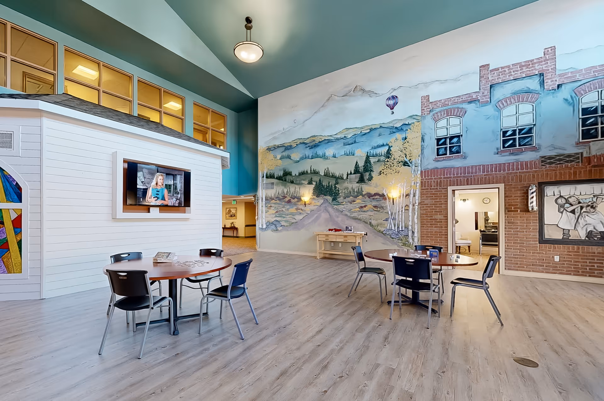 Spacious indoor common area with round tables and chairs, a large painted mural on one wall, and a TV mounted on a white shingled structure.
