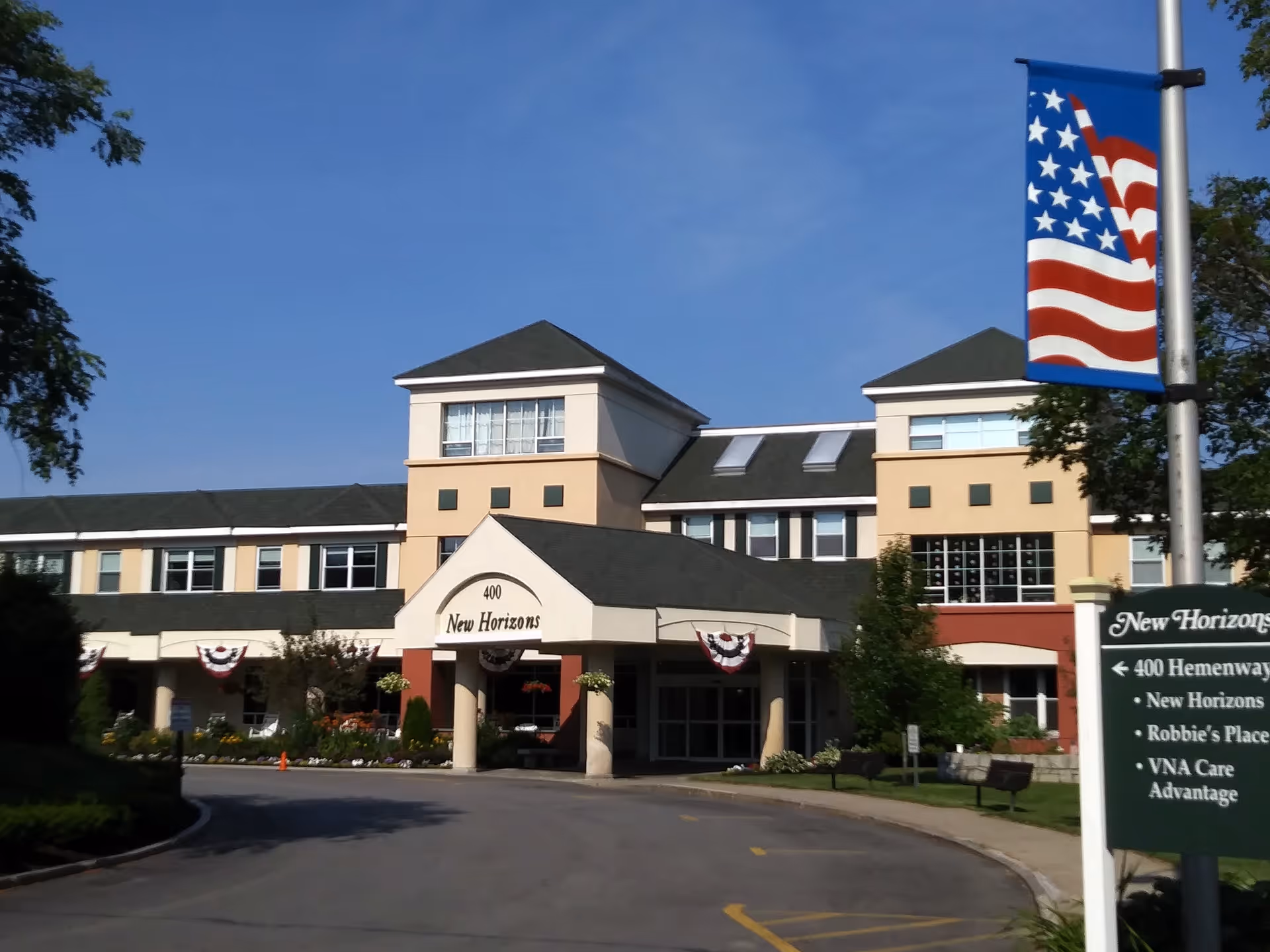 Front exterior view of the New Horizons at Marlborough senior living facility building with a covered entrance, American flag banner on a lamppost, and a sign listing facility locations.