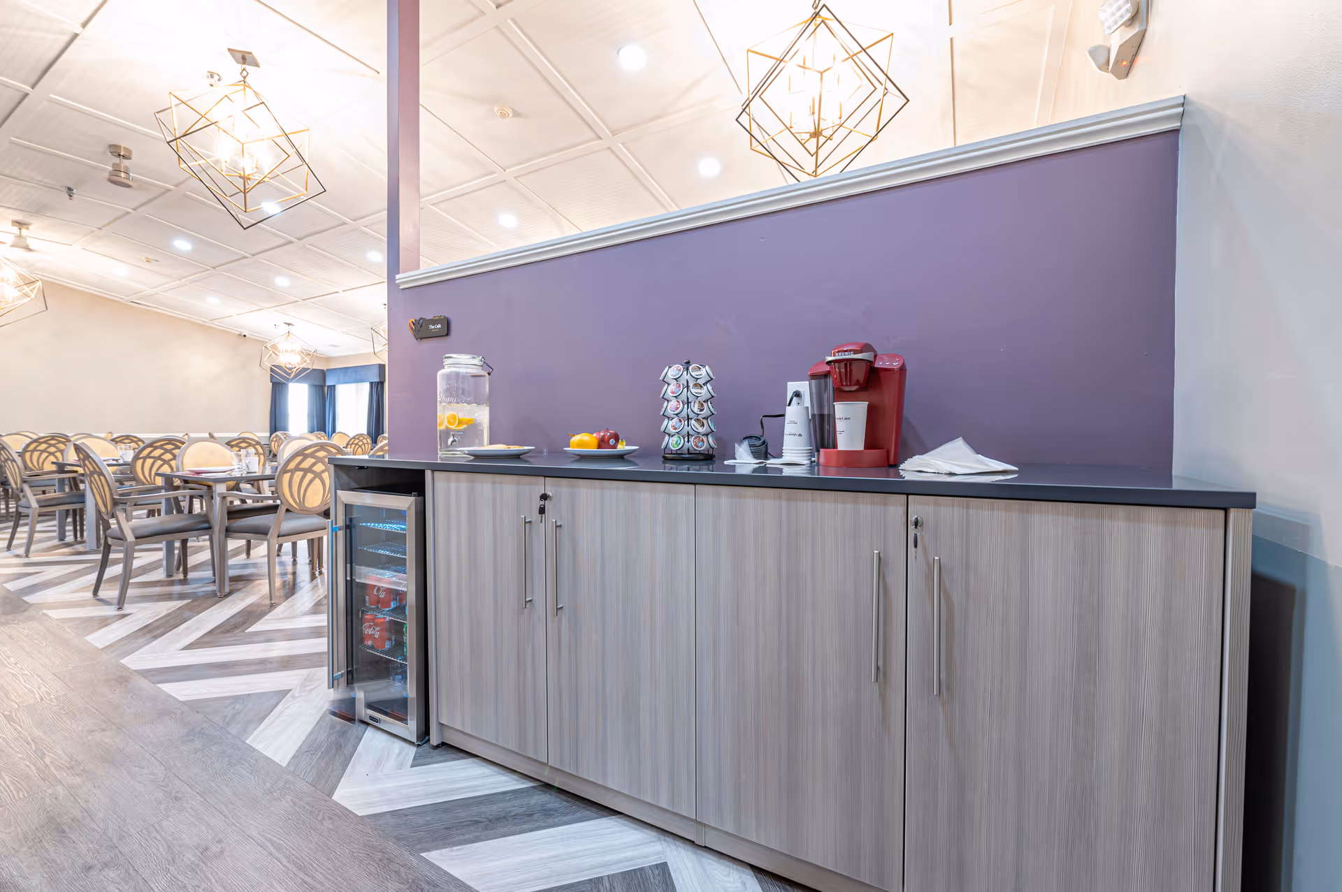 Interior view of a dining area in a senior living facility with multiple tables and chairs arranged neatly. In the foreground, there is a cabinet with a small refrigerator stocked with beverages, a water dispenser with lemon slices, a coffee maker, disposable cups, and a rotating rack of coffee pods. The wall behind the cabinet is painted purple, and the ceiling has modern geometric light fixtures.
