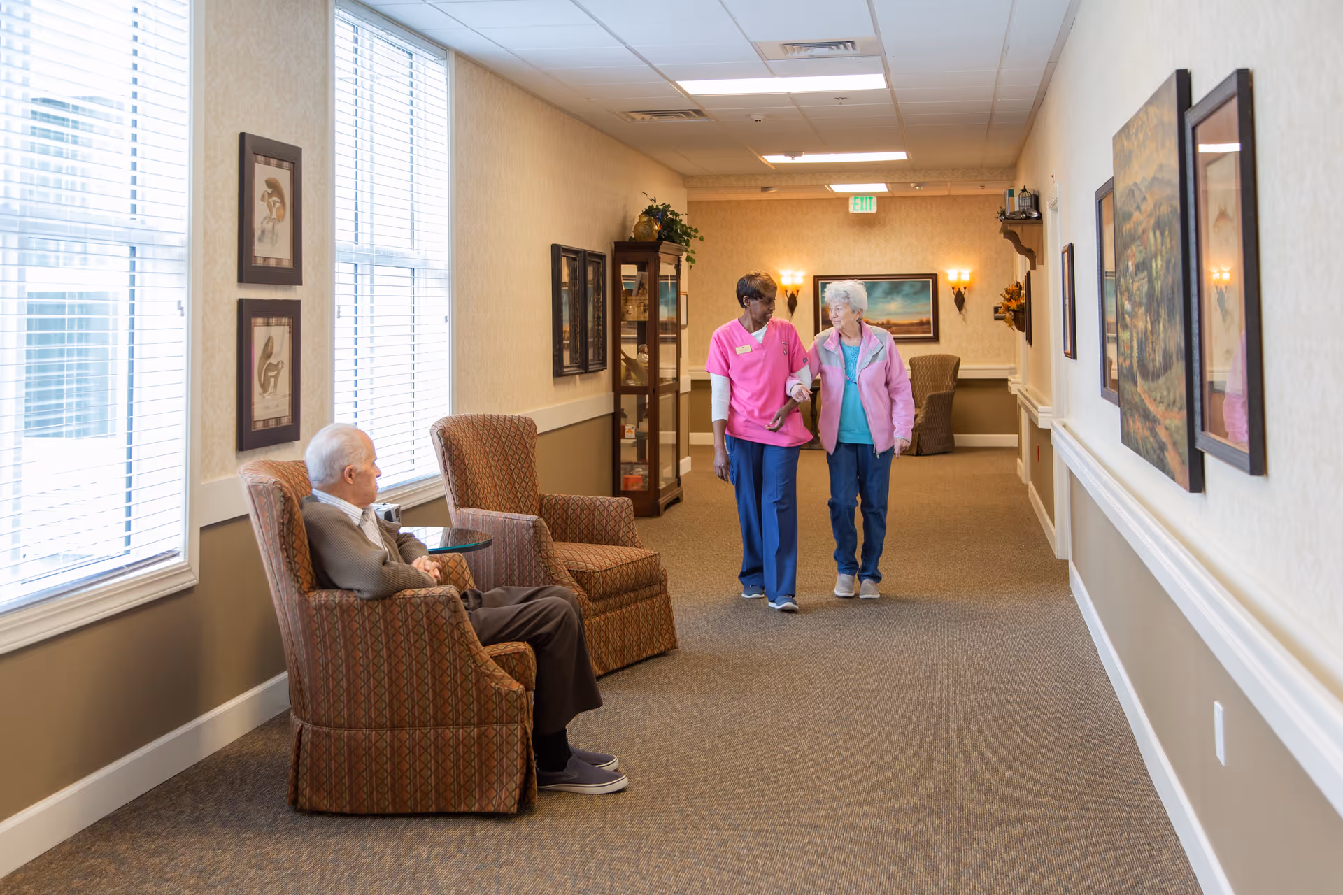 A hallway in an assisted living facility with two elderly individuals. One elderly man is seated in a patterned armchair near large windows with blinds, while an elderly woman walks arm-in-arm with a caregiver dressed in pink scrubs. The hallway is decorated with framed artwork, wall sconces, and a display cabinet.