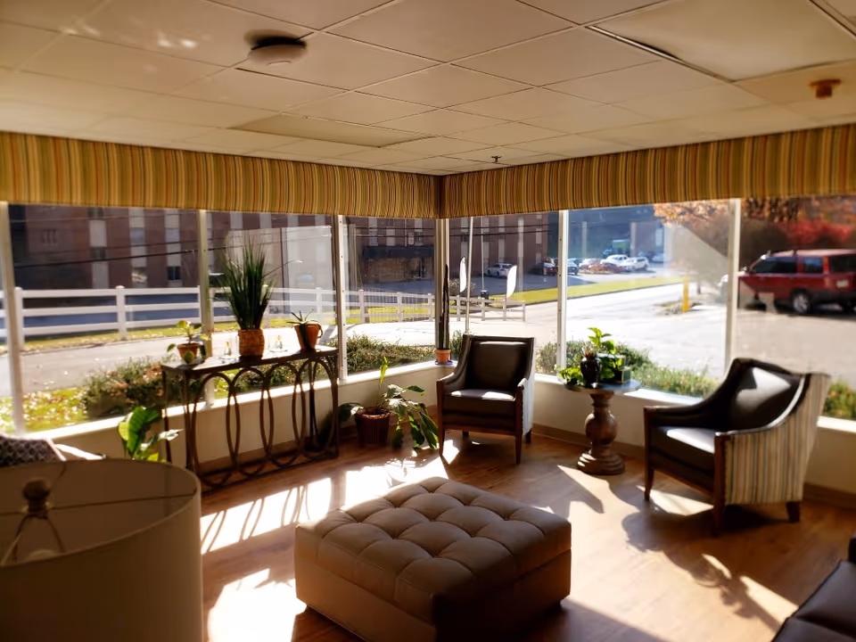 A sunlit sitting area with large windows showing an outside view of a street and parked cars. The room has two armchairs, a cushioned ottoman, a small round table with plants, and a console table with more plants. The ceiling has a grid pattern, and the windows have striped valances.
