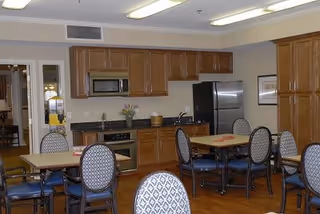 A dining area with several tables and chairs featuring patterned upholstery. The room includes a kitchen area with wooden cabinets, a microwave, an oven, and a stainless steel refrigerator. The floor is wooden, and the walls are light-colored with a framed picture visible.