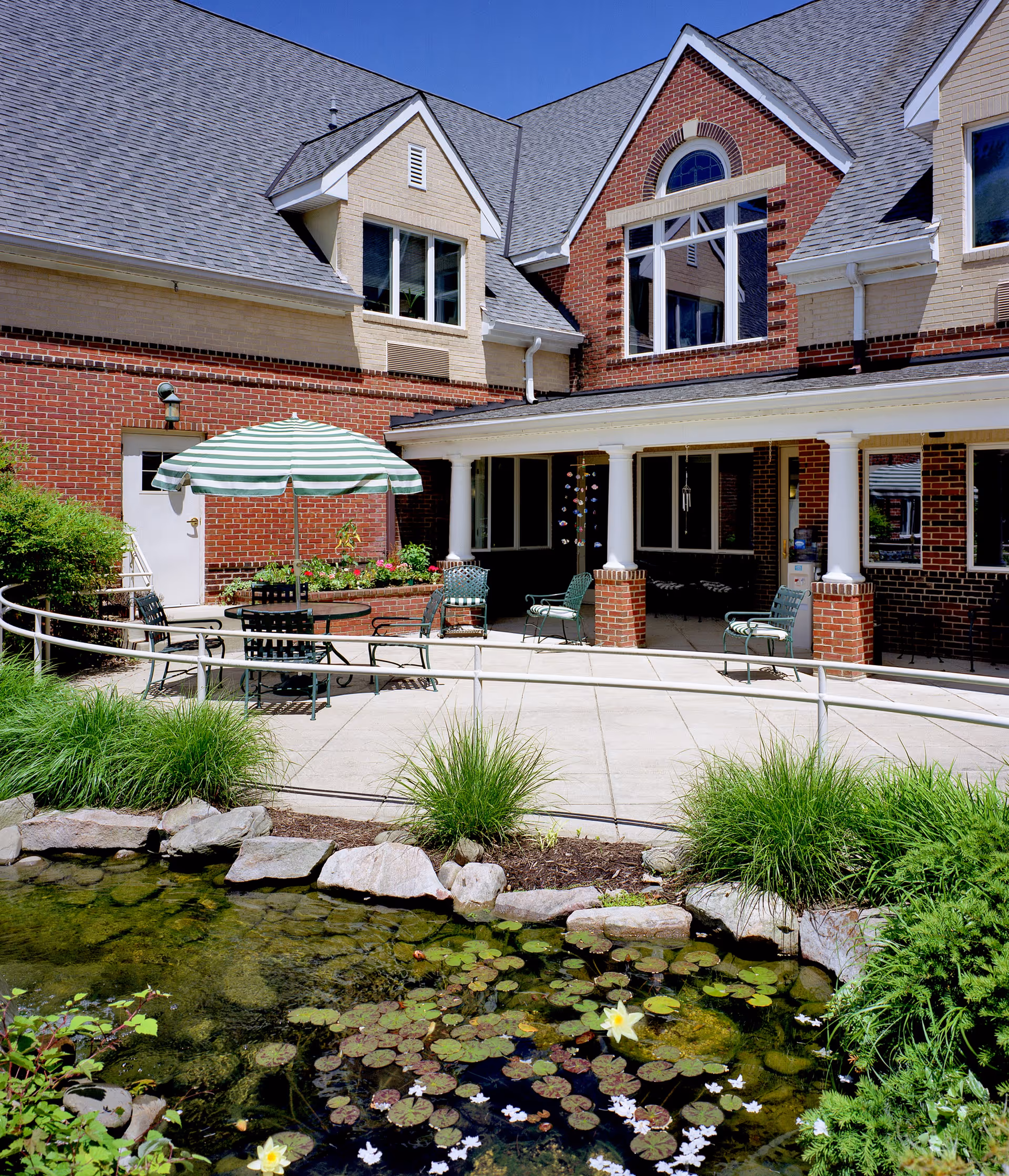 Outdoor courtyard with patio seating and a striped umbrella beside a lily pond in front of a brick senior living building.