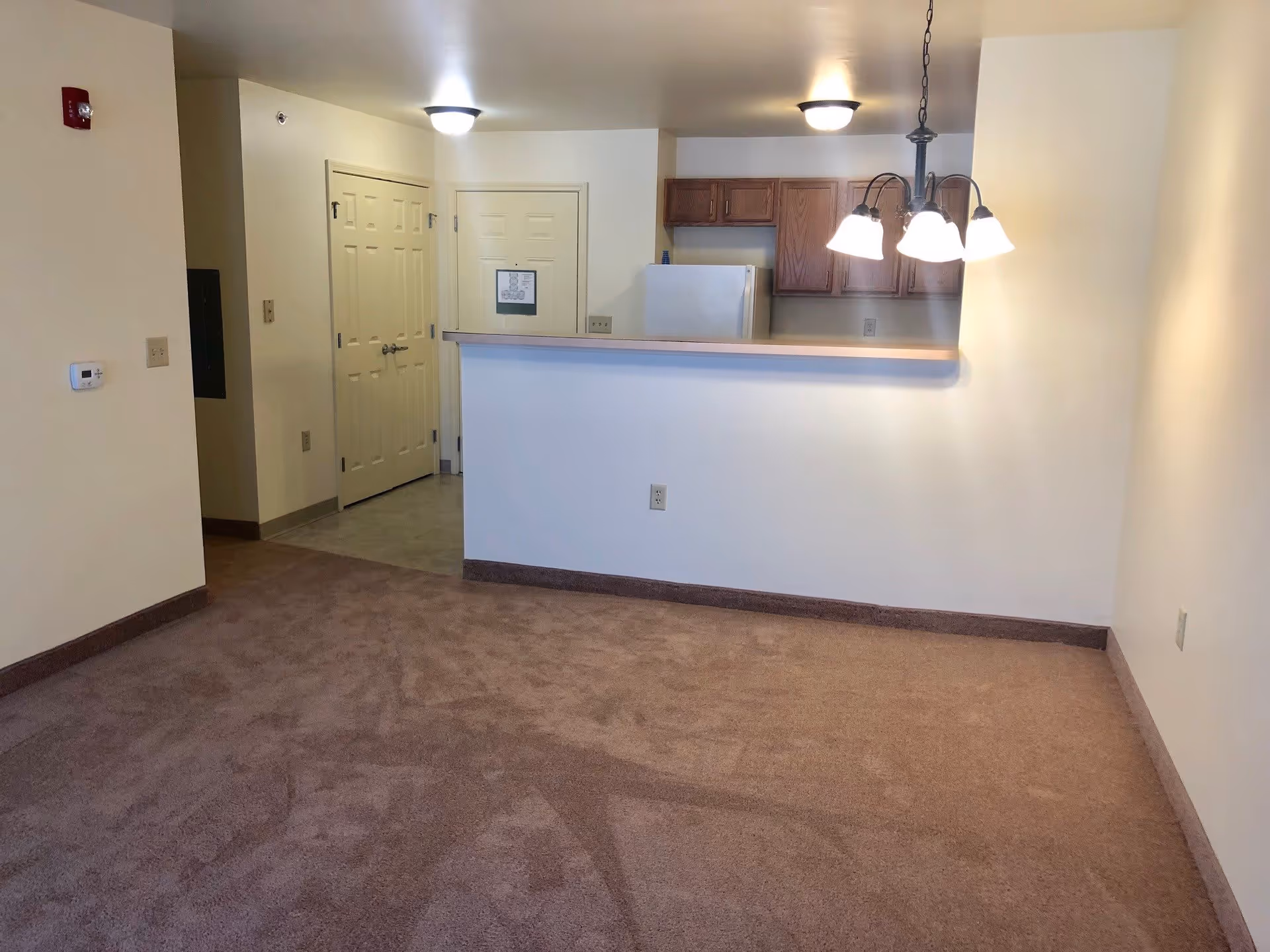 Empty carpeted living area with a counter-style pass-through to a small kitchen, hanging light fixture, and white walls.